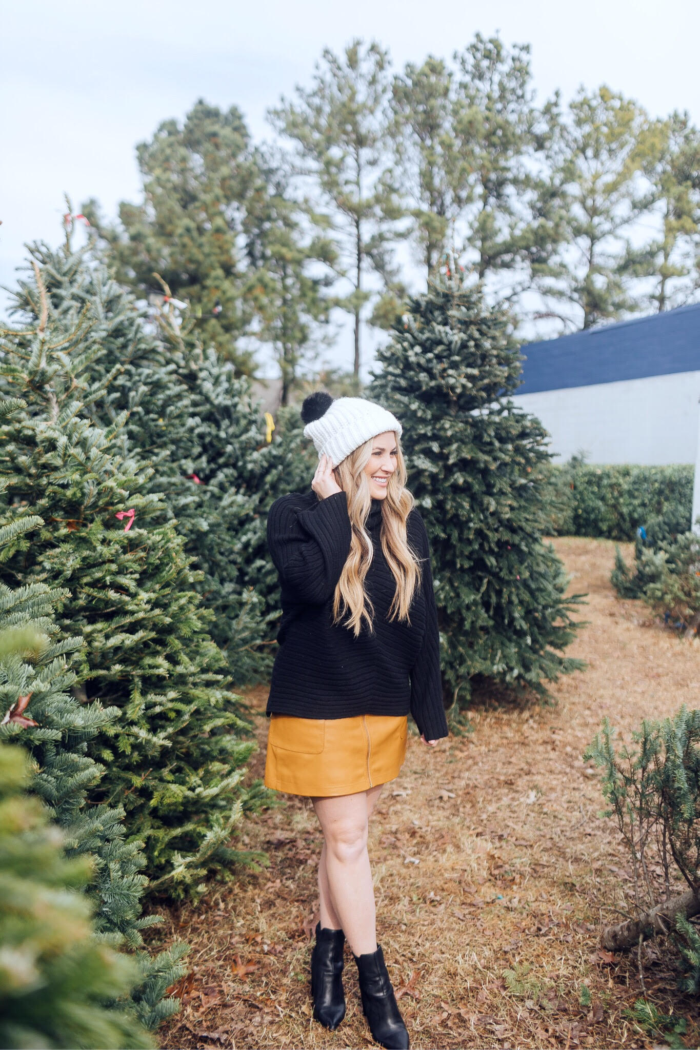 Winter accessories featured by top US fashion blog, Walking in Memphis in High Heels: image of a blonde woman walking in a Christmas tree farm wearing a Free People mini skirt, Melroseand Market sweater, Rebecca Minkoff pompom beanie, Sole Society Sock Bootie