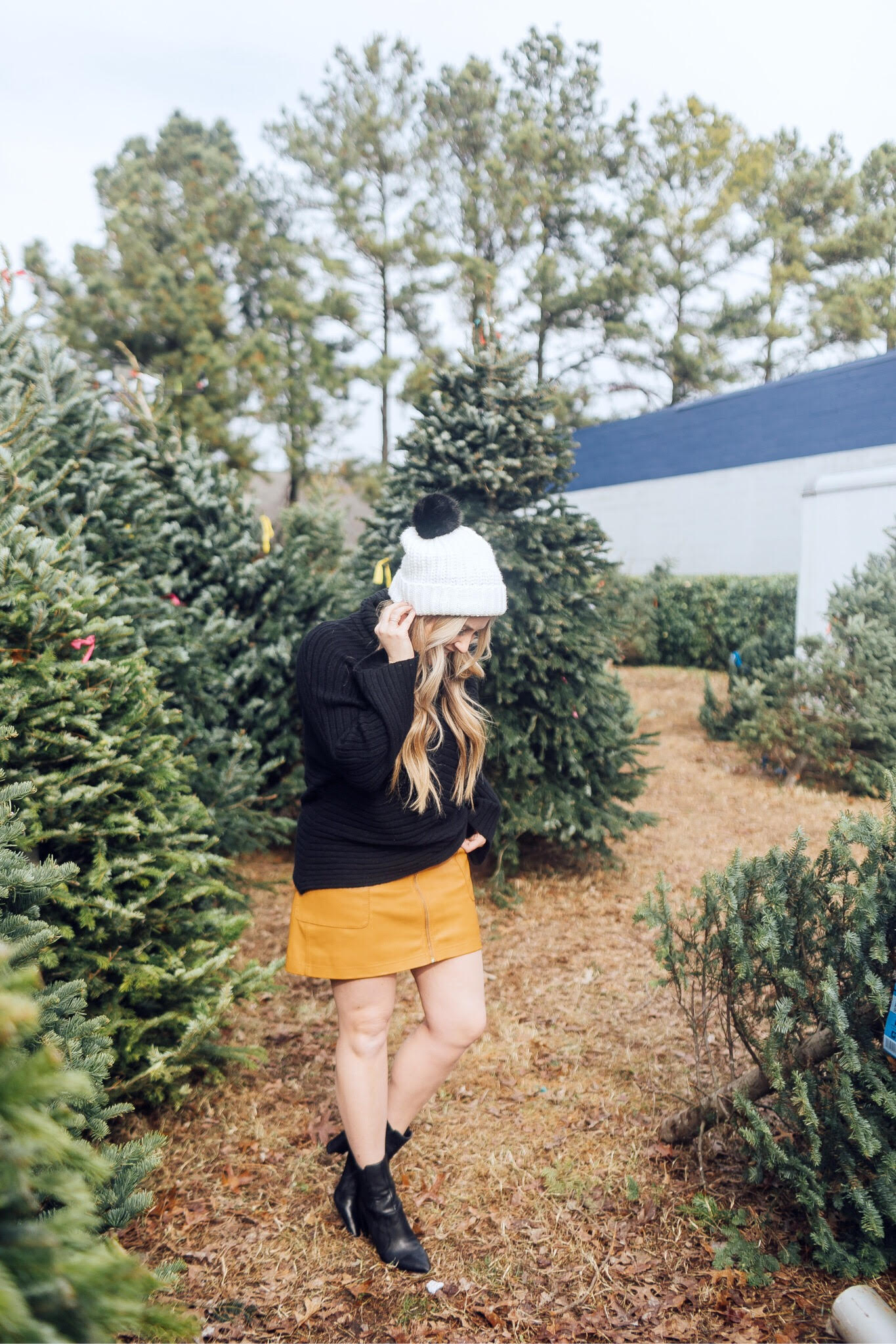Winter accessories featured by top US fashion blog, Walking in Memphis in High Heels: image of a blonde woman walking in a Christmas tree farm wearing a Free People mini skirt, Melroseand Market sweater, Rebecca Minkoff pompom beanie, Sole Society Sock Bootie